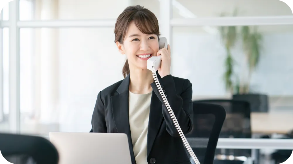 オフィスで電話対応をする笑顔の女性スタッフ。パソコンの前で丁寧に応対している様子。